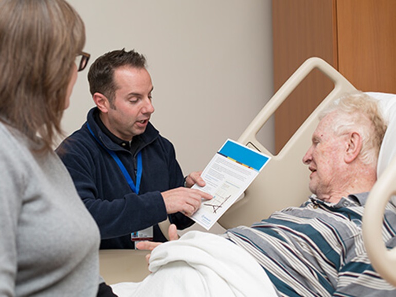 Male nurse showing a medical form to a senior male patient lying in hospital bed.