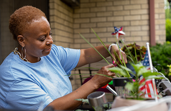 A woman is on her porch watering her plants with a few American flags in the baclground