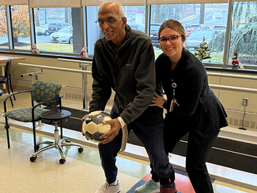 Srini Muralidhar holds onto a medicine ball and lunges forward as a therapist holds him by the waist. Behind them are the windows and equipment of the Helen M. Simpson Rehabilitation Hospital gym
