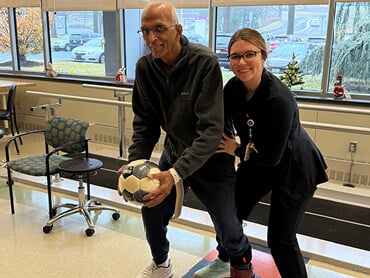 Srini Muralidhar holds onto a medicine ball and lunges forward as a therapist holds him by the waist. Behind them are the windows and equipment of the Helen M. Simpson Rehabilitation Hospital gym